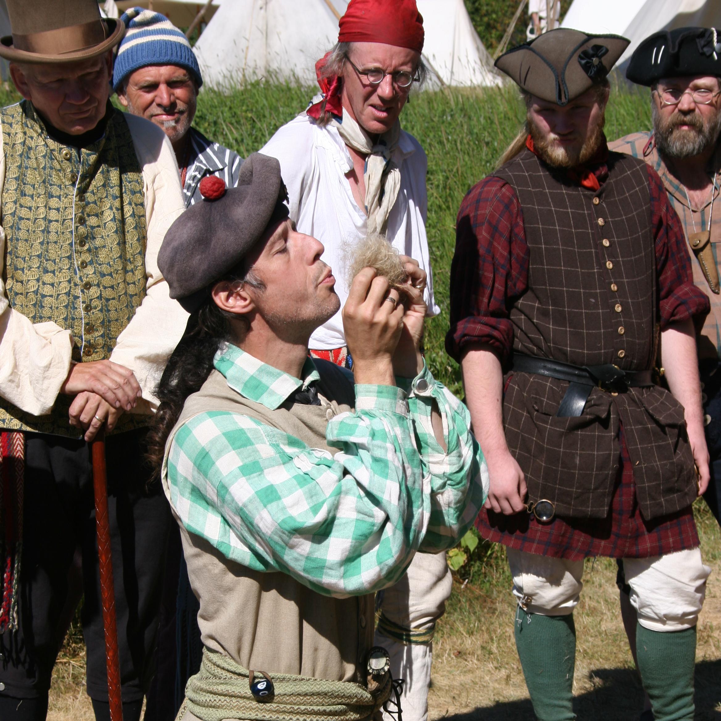 People watching a kneeling man in historic clothing blowing on some fluff.