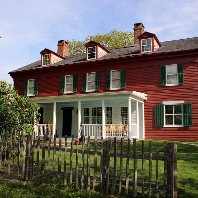 A large red house, 2 stories tall, with a sloped black roof, green shutters, and a white porch. 