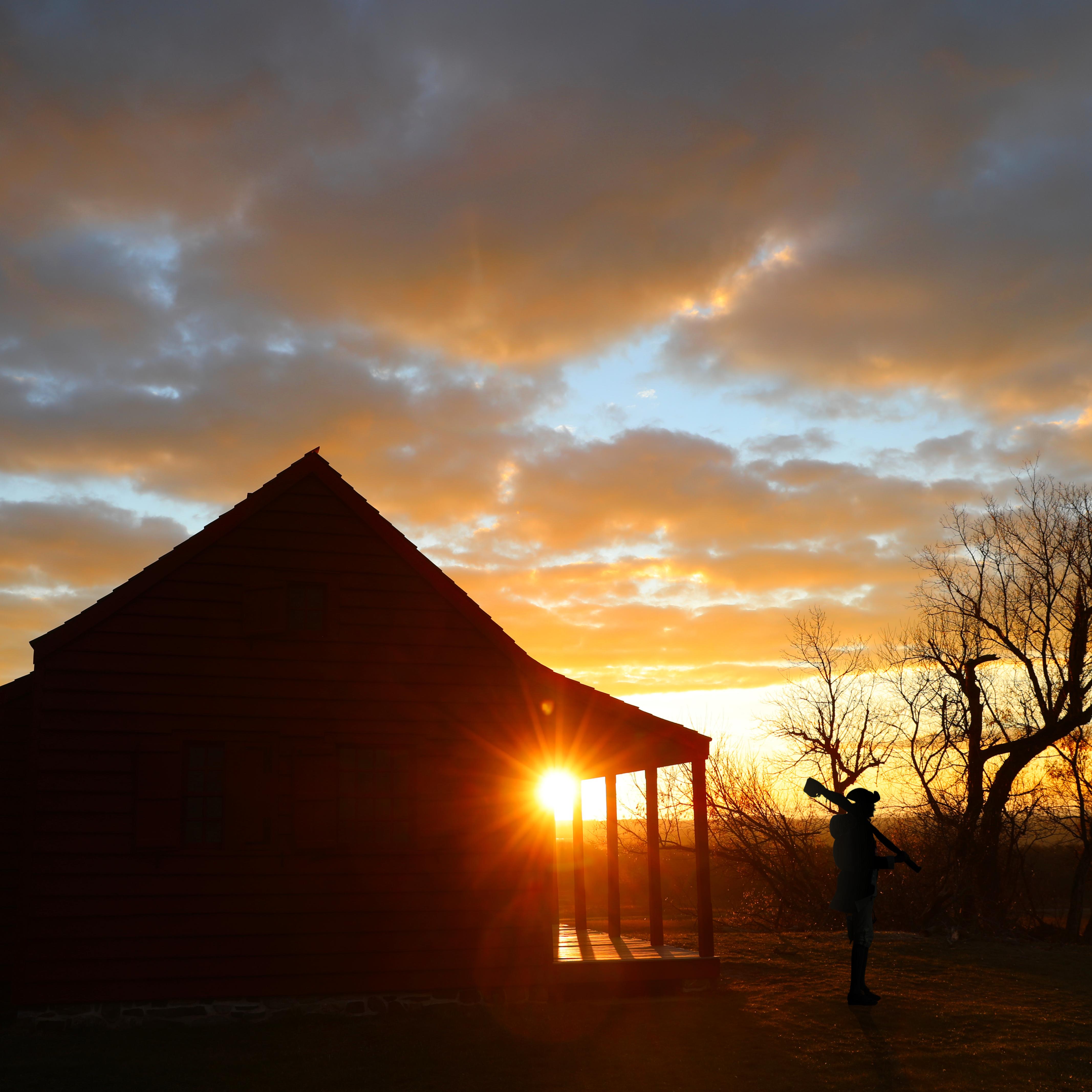 A man in 18th-century style clothing carrying a musket and a farmhouse are silhouetted by sunrise.