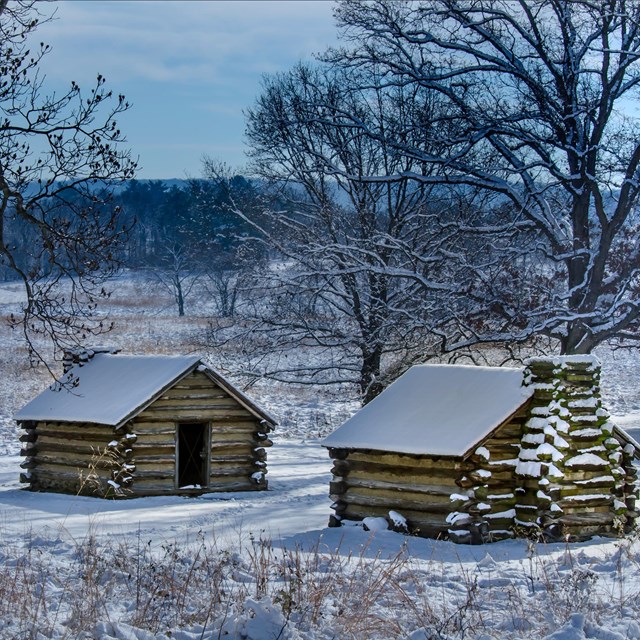 2 small wooden cabins with large chimneys. They are covered by a dusting of snow in an open field. 