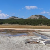 A thermal area sits in a meadow in front of rolling hills.