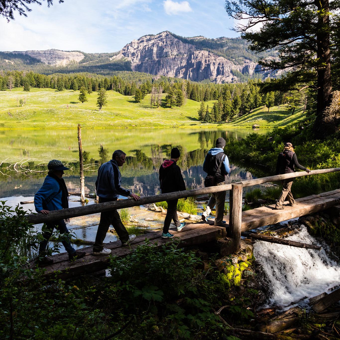 Hikers walk across a bridge at the outlet of a small lake surrounded by rolling hills.