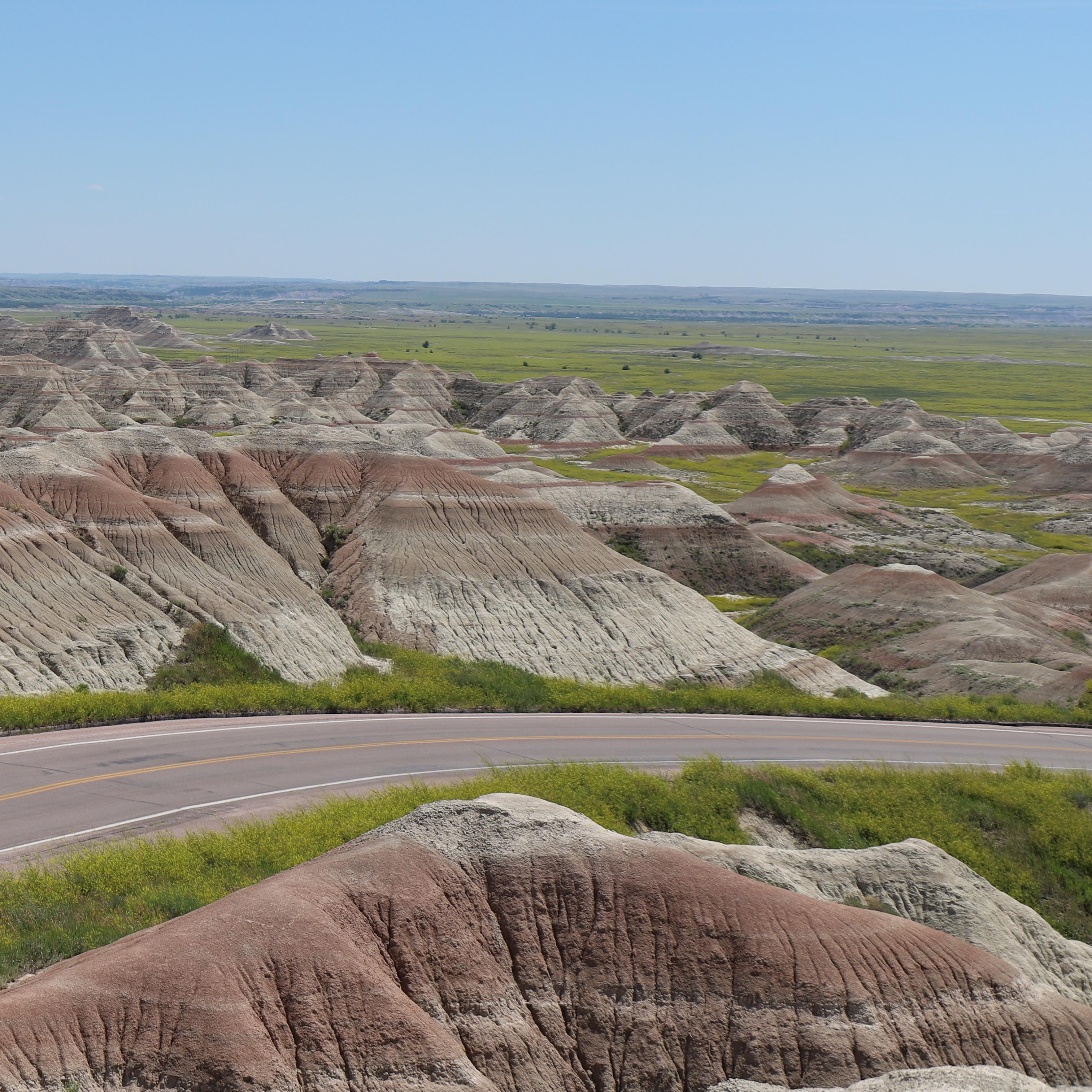 a paved road descends into a prairie landscape with buttes in the background.