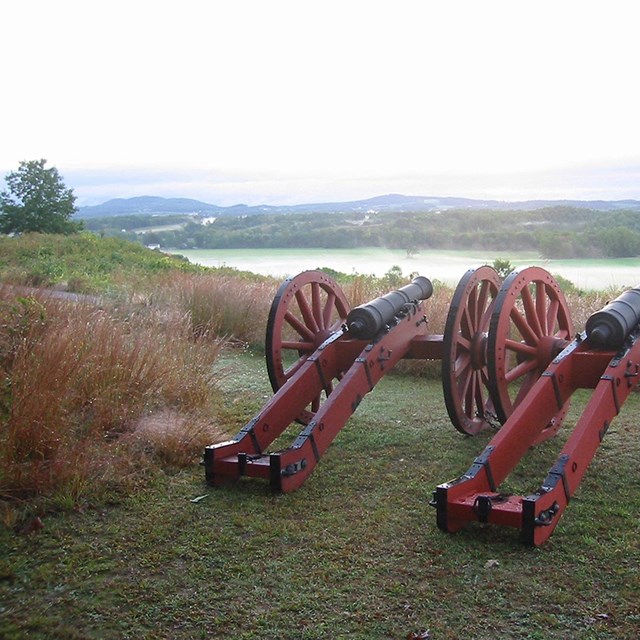 2 red field cannons sitting on a bluff overlooking the Hudson River. It cuts a green valley in half.
