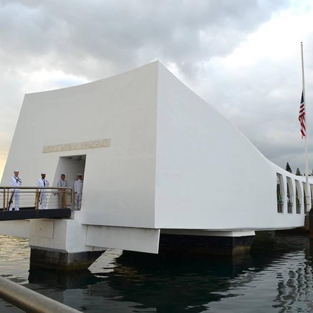 A long white memorial sits over the water, people are entering the memorial, a flag is at 1/2 staff