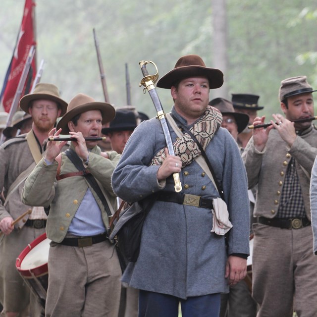 A formation of men walking in various grey wool uniforms. At the lead, a man with a sword. 