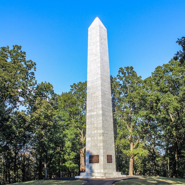 A white stone, spire monument juts upward from green grass to a blue sky.