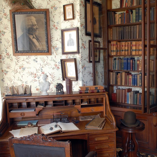 A portrait of Frederick Douglass hang on a wall in a library filled with books and a desk.