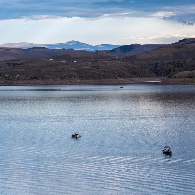 A large blue reservoir is surrounded by mesas. Three boats are on the water in the distance.