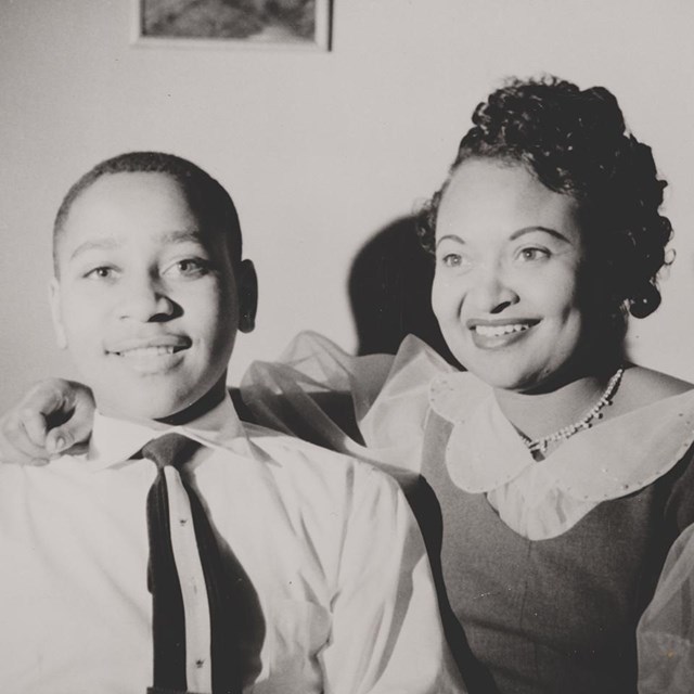 A Black woman and her son smile while sitting on a couch. The woman has one arm around his shoulder.