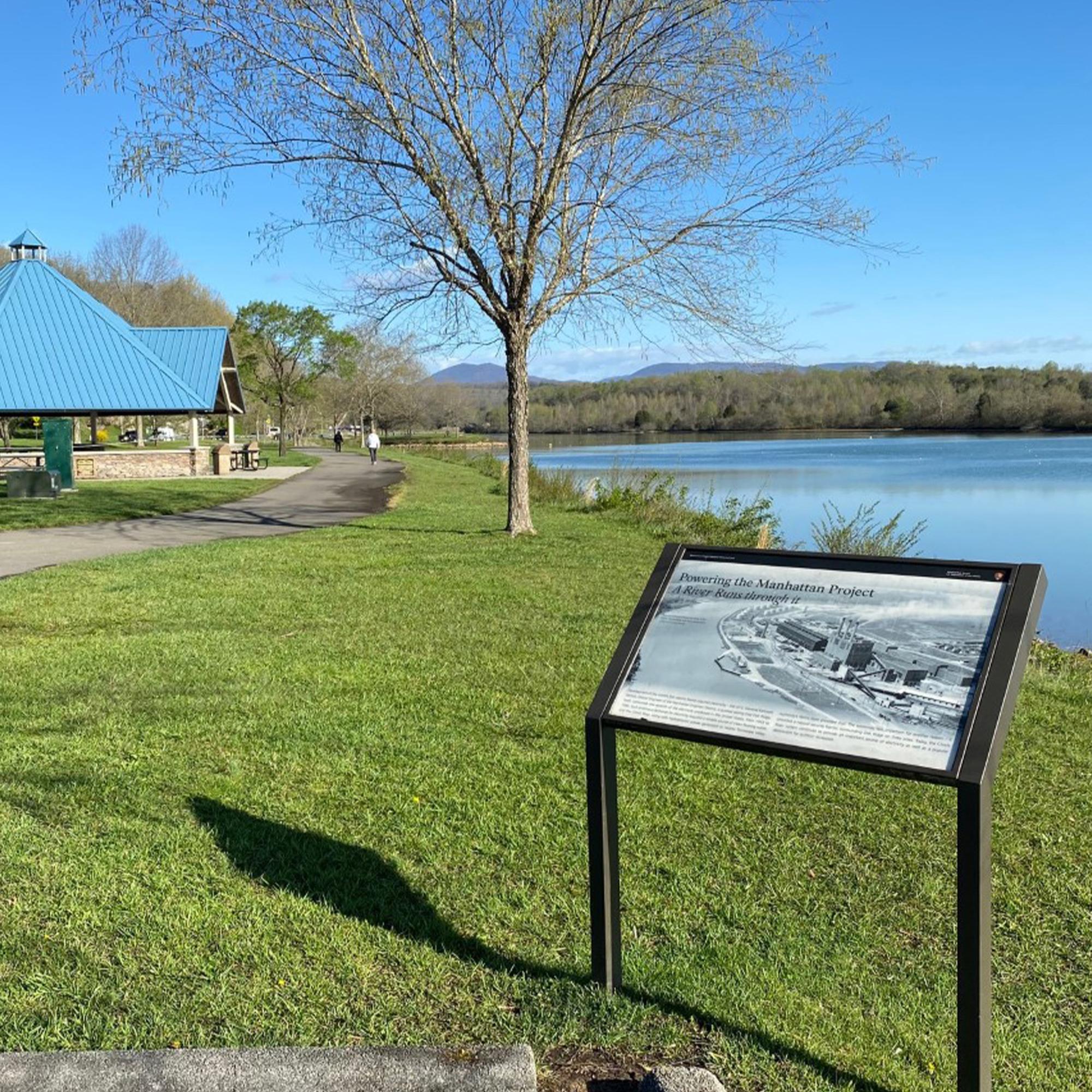 An historical marker on the bank of a calm river with a paved trail and pavilion in the background. 
