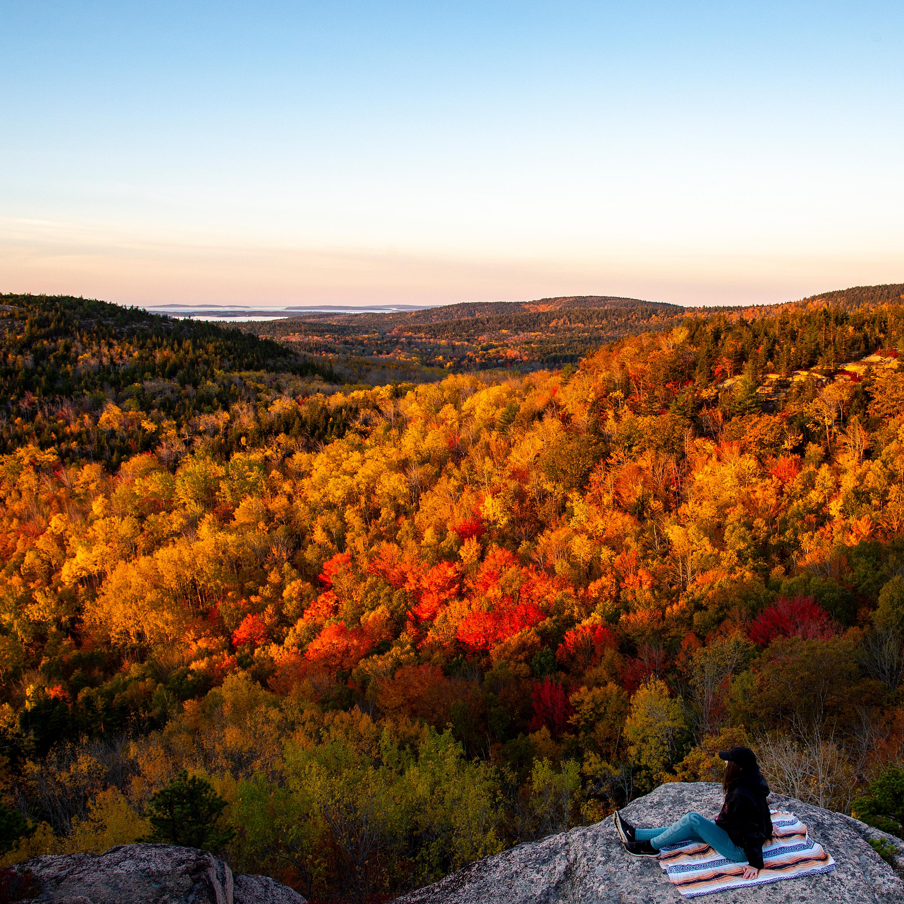 A person sits on a rock ledge looking out over sunny hills with colorful fall foliage
