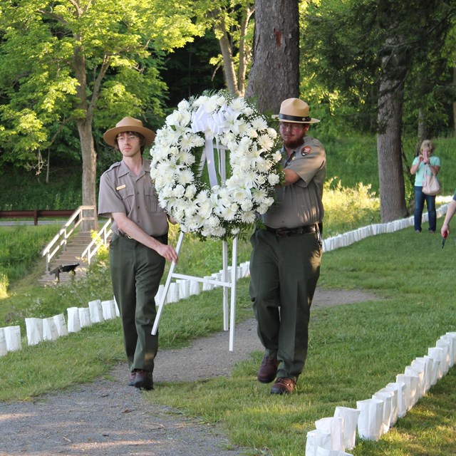 2 park rangers walk down a grassy path with a large white wreath carried between them. 
