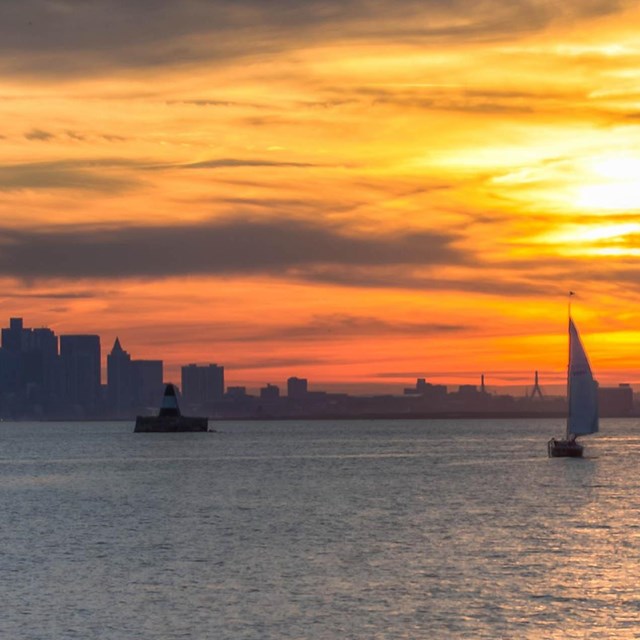 A fiery sunset over water. Reds, yellows, reds, & blues fade to grey of a city skyline and sailboat.