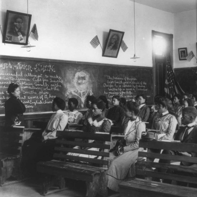 An all African American class look at a teacher standing by a chalkboard; B&W Photo