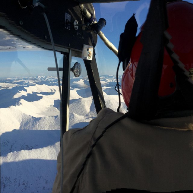 A pilot with a red helmet on, flying a plane high above snowy terrain