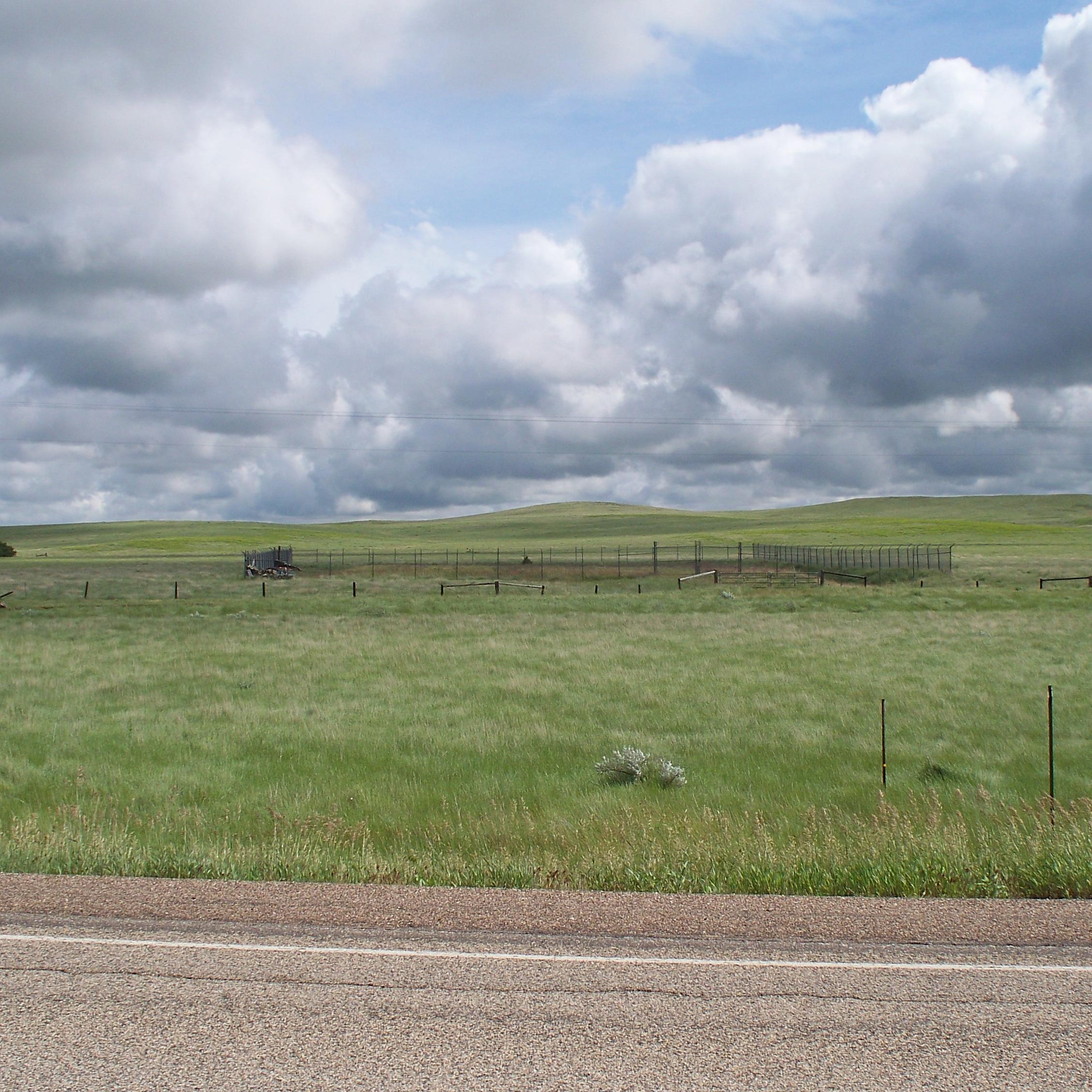 View from a highway into the prairie; in the foreground a rectangular fence breaks up a field.