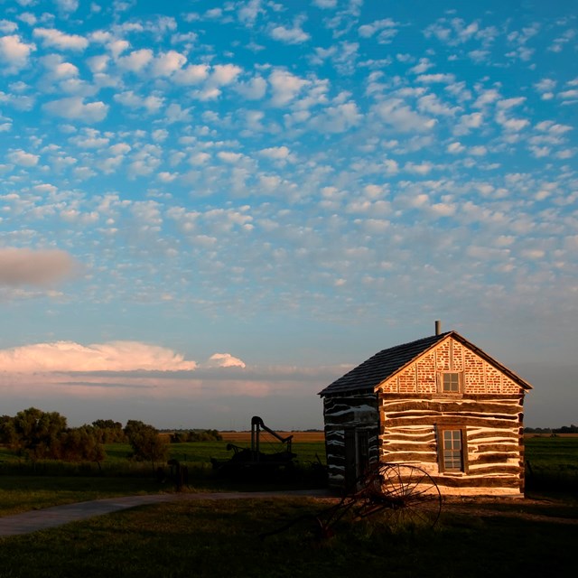 A square log cabin sits to the left of an expanse of prairie. 