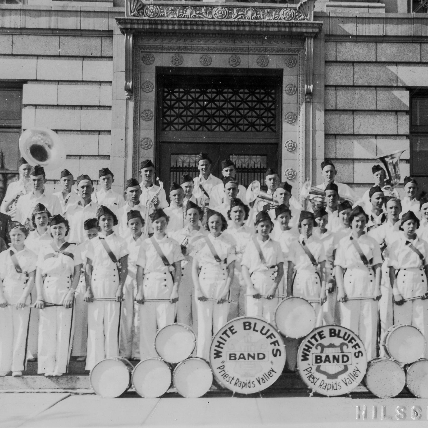 Historic photo of a school band in uniforms standing in front of a  brick building