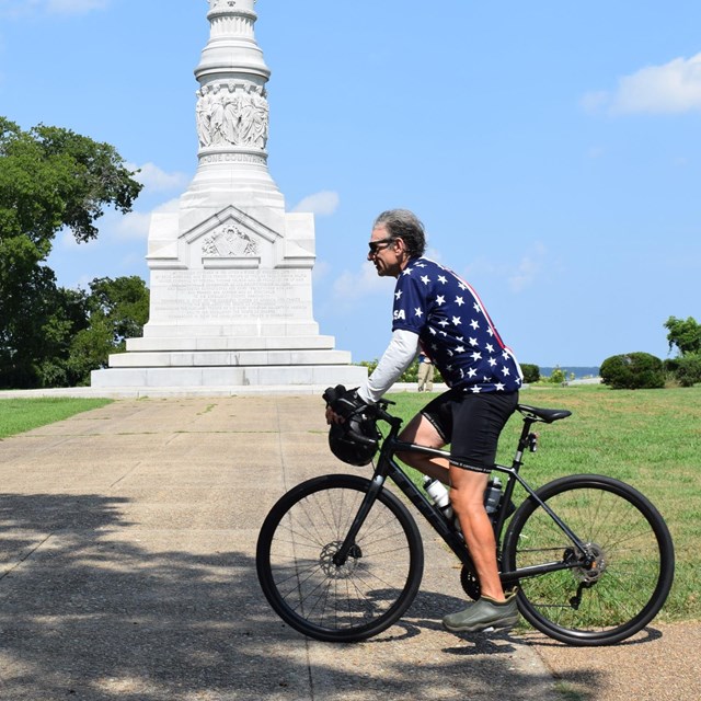 a man in a red, white and blue jersey rides a bicycle in front of a granite monument