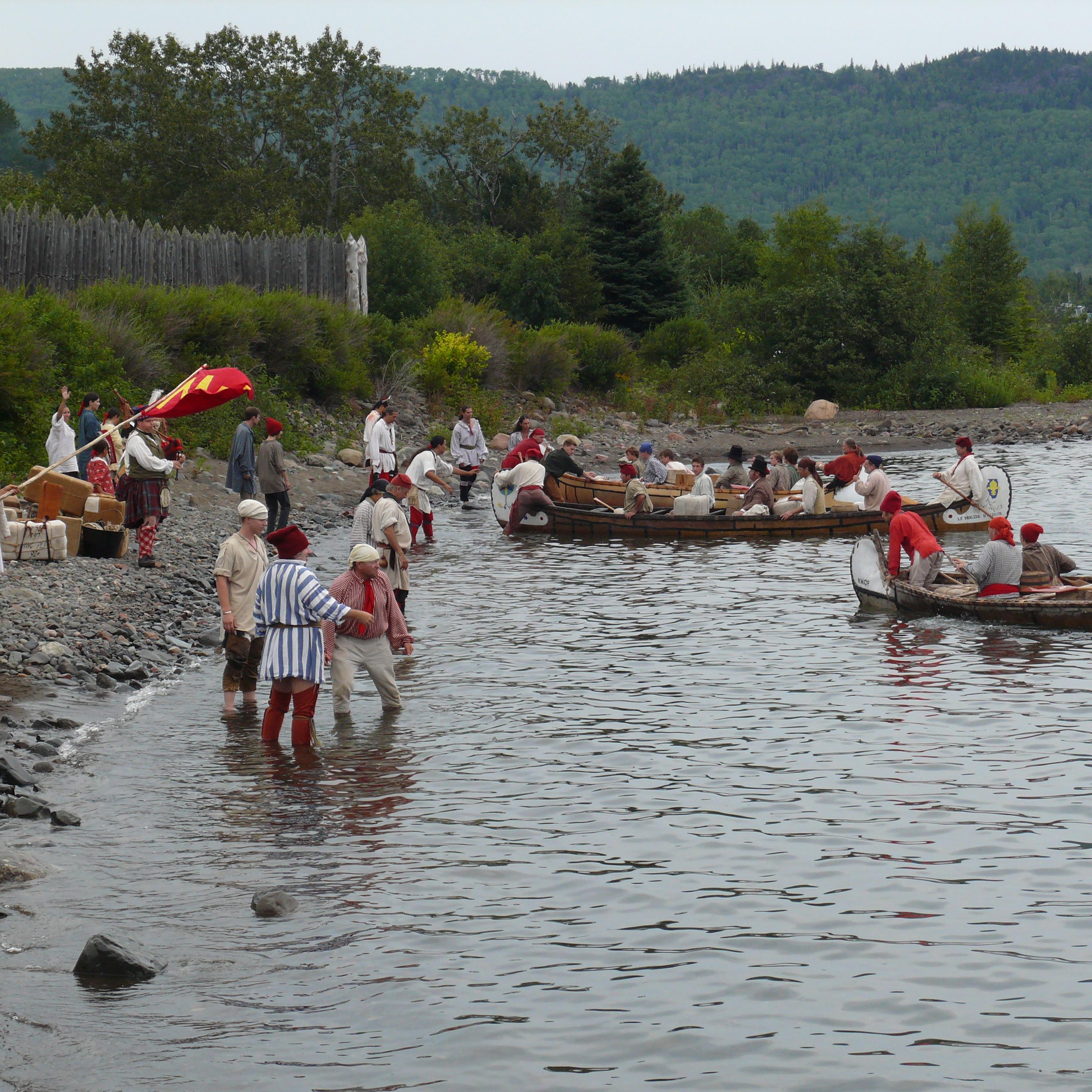 Canoes and people wearing historic clothing at the edge of a lake, next to a stockade.