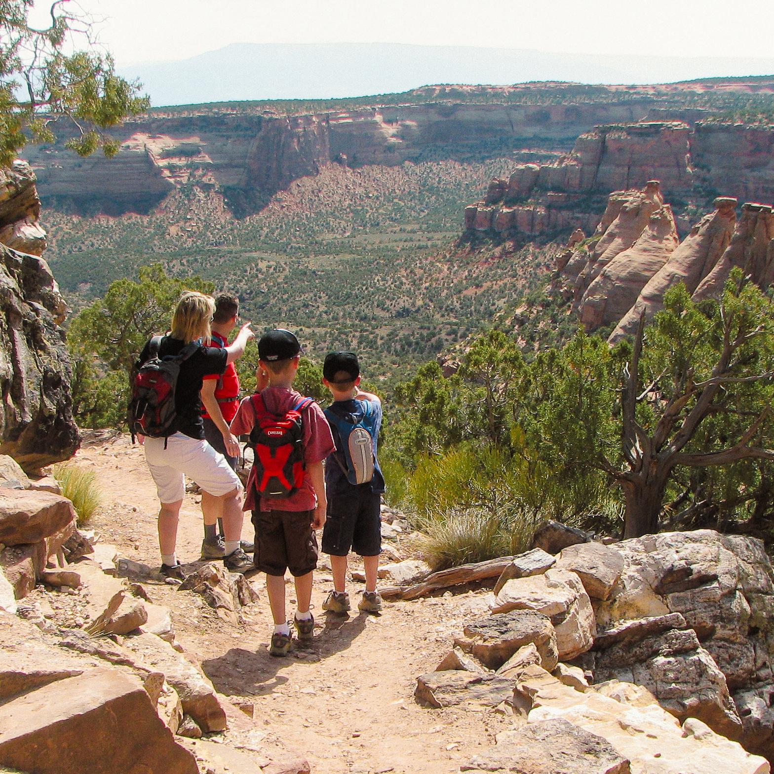 two adults and two children stand on dirt path looking out towards wide open canyons