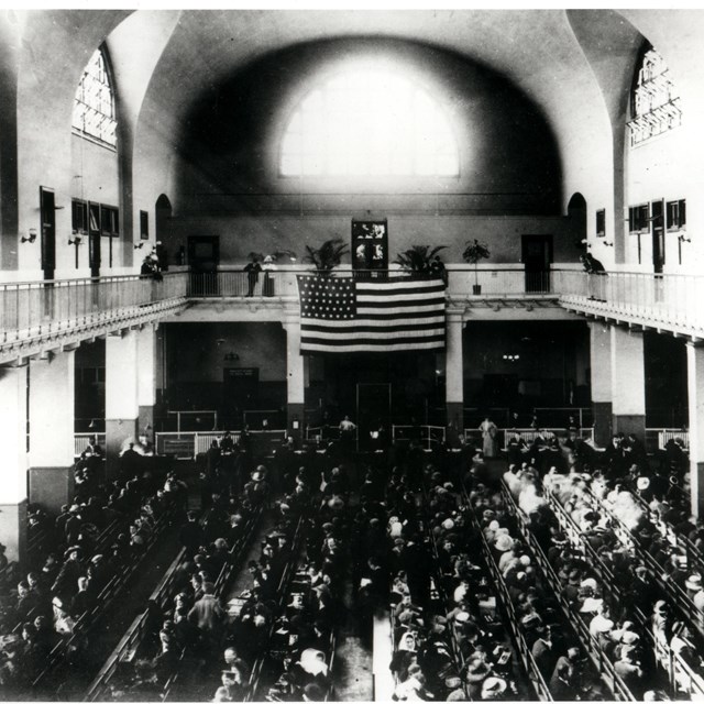 A black and white image of a large hall. An American flag hangs over a throng of people on the floor