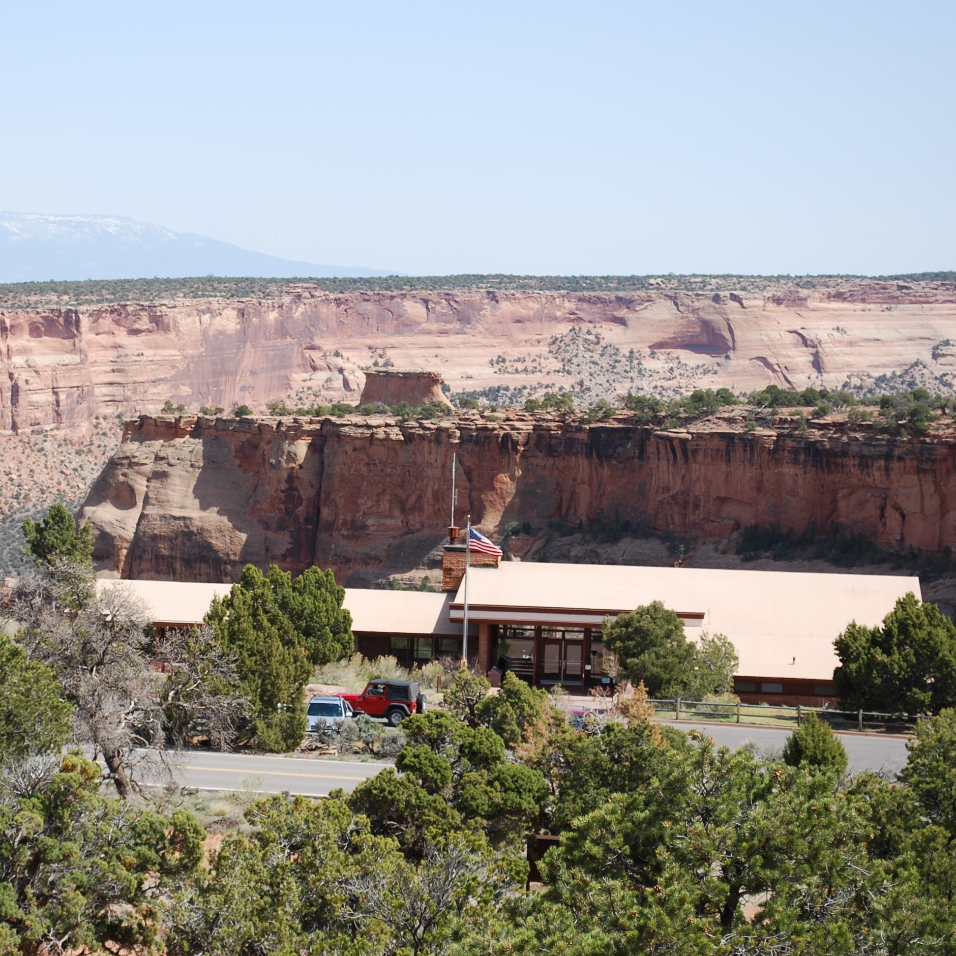 A low, mostly flat building with a flagpole and shrubs, standing before a desert canyon