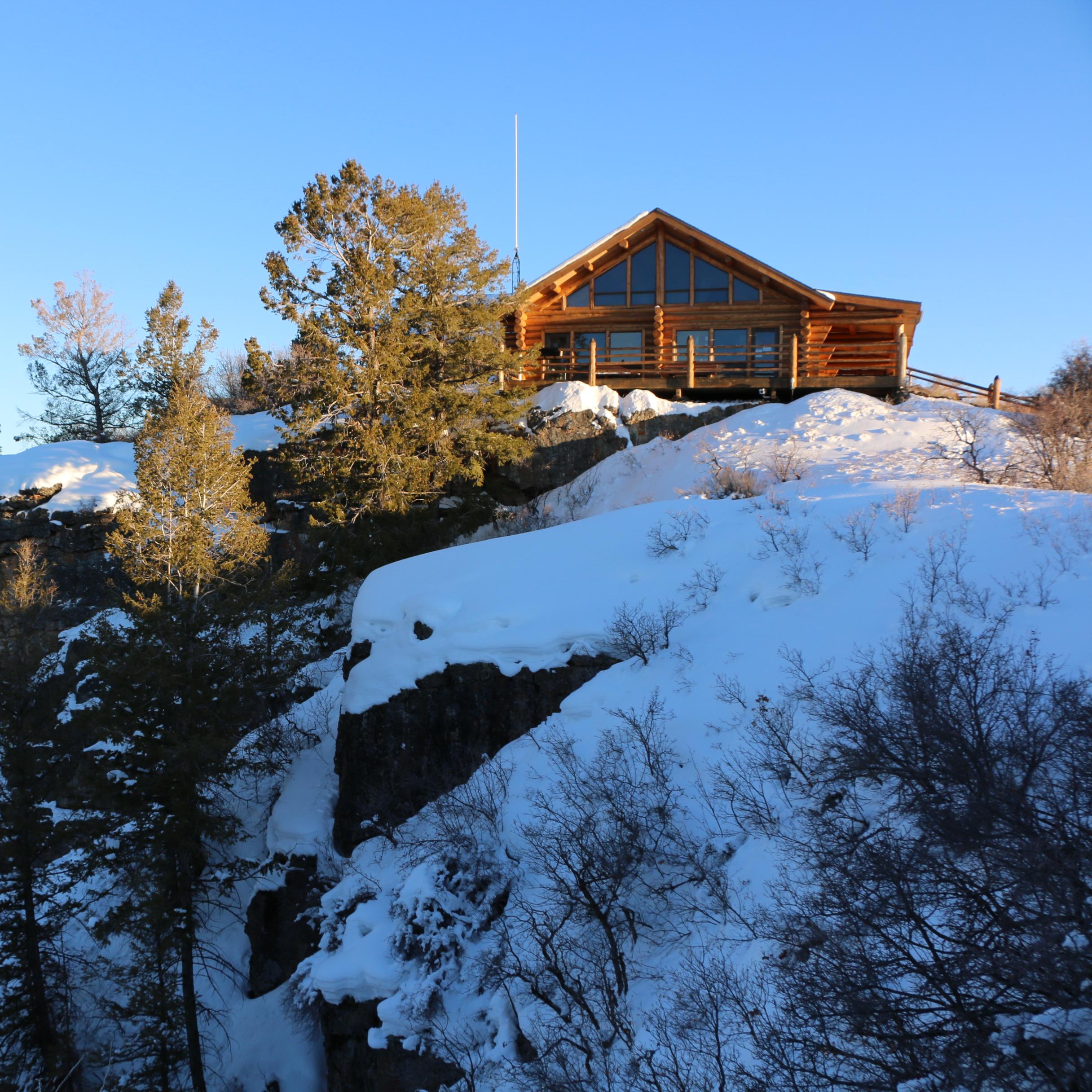 A log structure with large windows sits on the edge of a steep, snowy canyon wall.