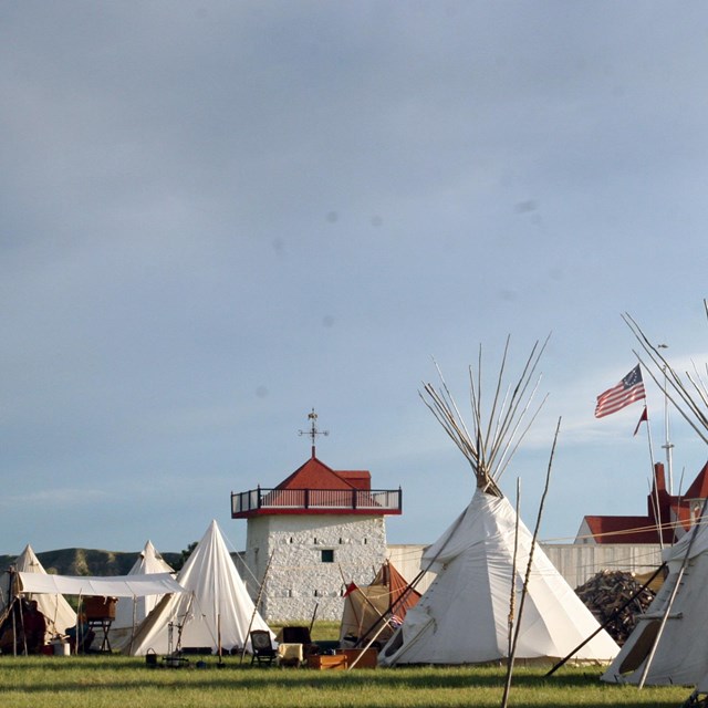 Landscape photograph. Canvas tipis stand tall in front of a red roofed whitewashed stone bastion. 