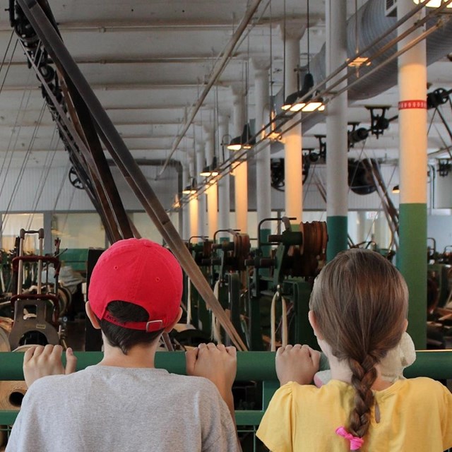 Two young visitors peer over the rail at the looms in the 1910s working weave room at the Boott Mill