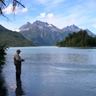 Fisherman with waders standing with fishing pole in lake with mountains in background