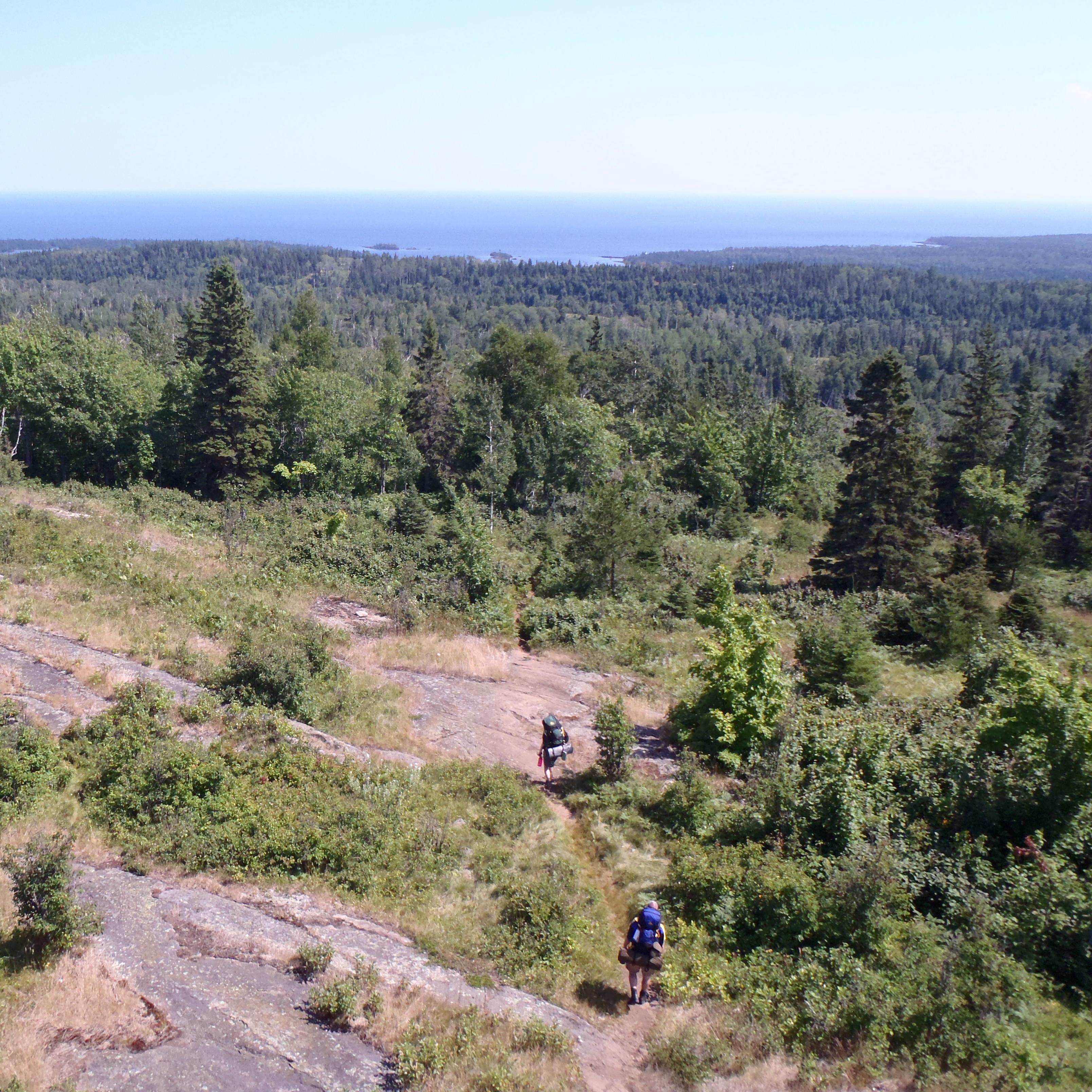 Two people with backpacks walk down a trail on a ridge surrounded by forest. 