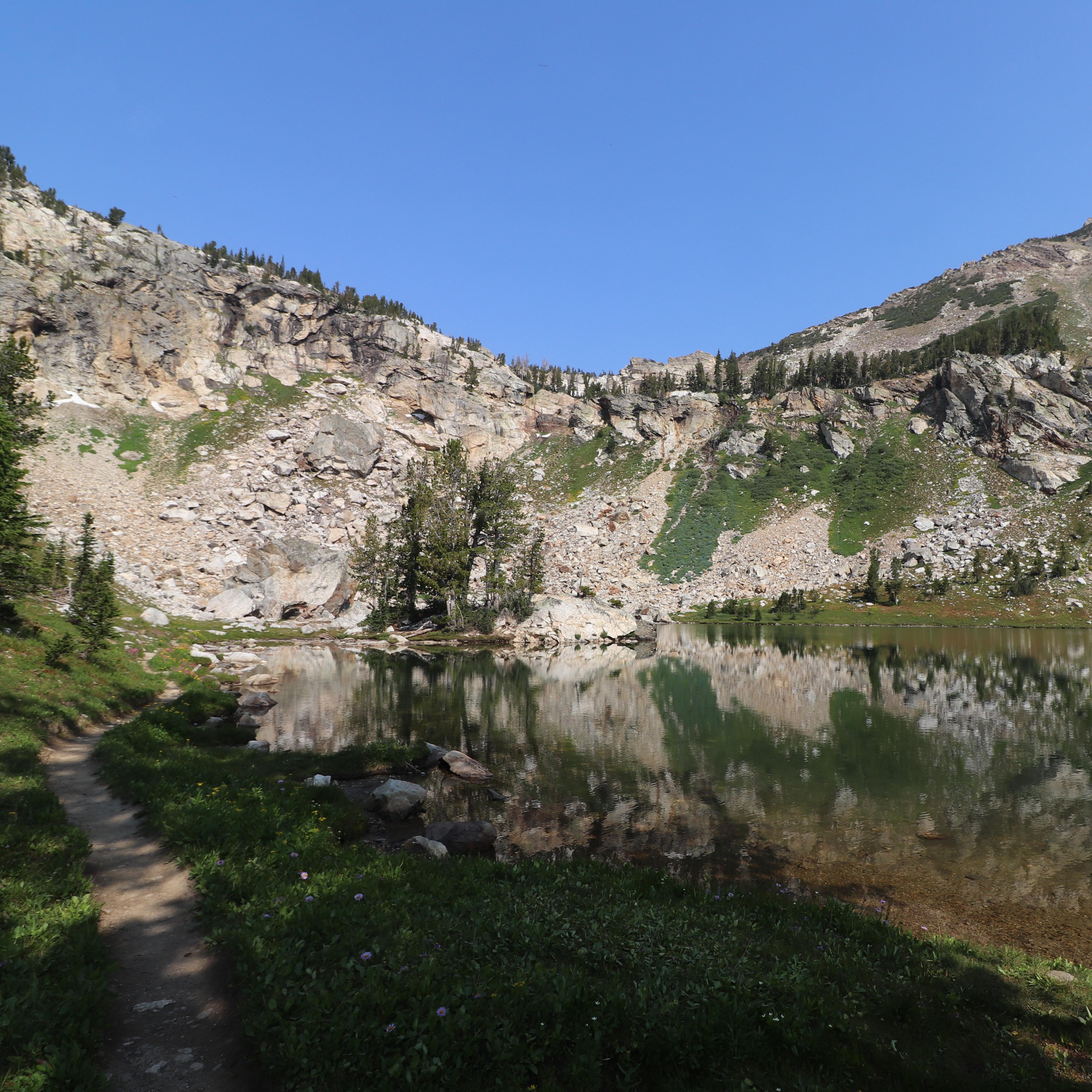 An alpine lake sits at the base of a rocky cliff surrounded by green vegetation.