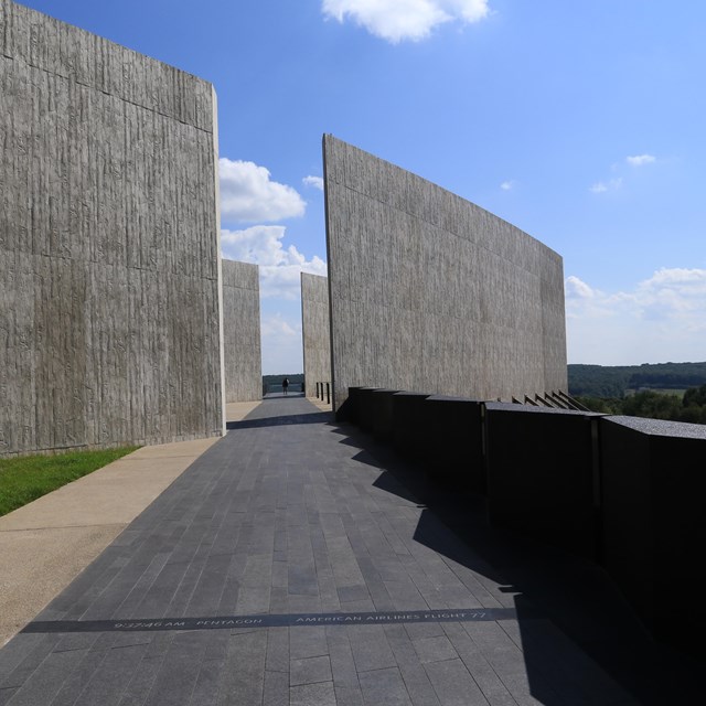 A black stone path through a large cement grey wall. You can see the blue sky in the opening.