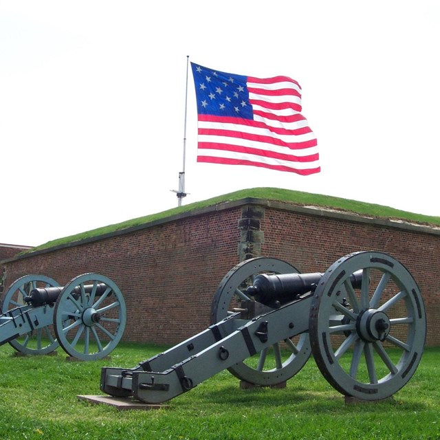 A large American flag waves over the wall of a large brick building. 2 grey cannons face the wall.