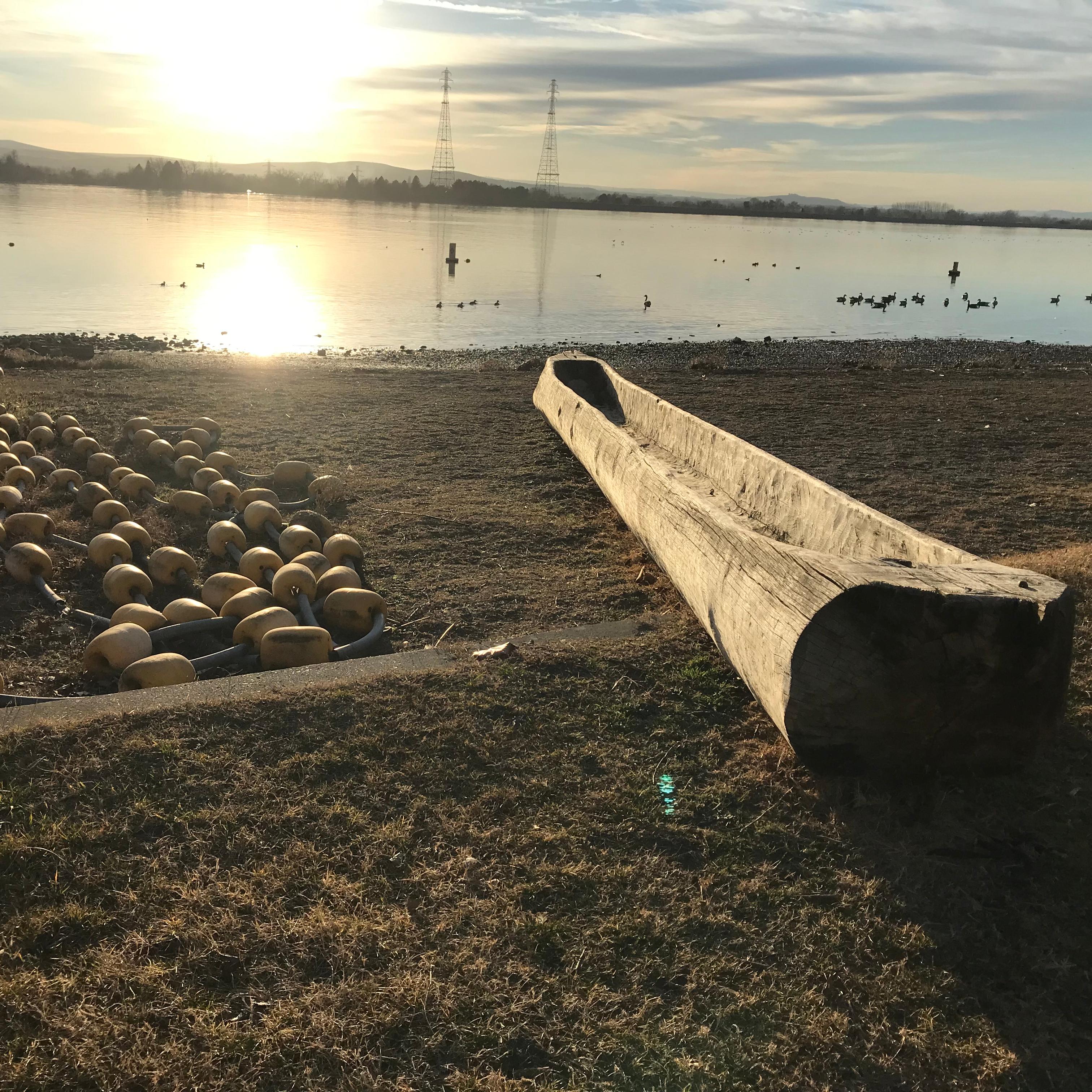 A color photo  of a large body of water with distant shores. There is a log dugout into a canoe.