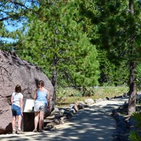 A girl and a woman stand next to a boulder and interpretive sign on a walkway.