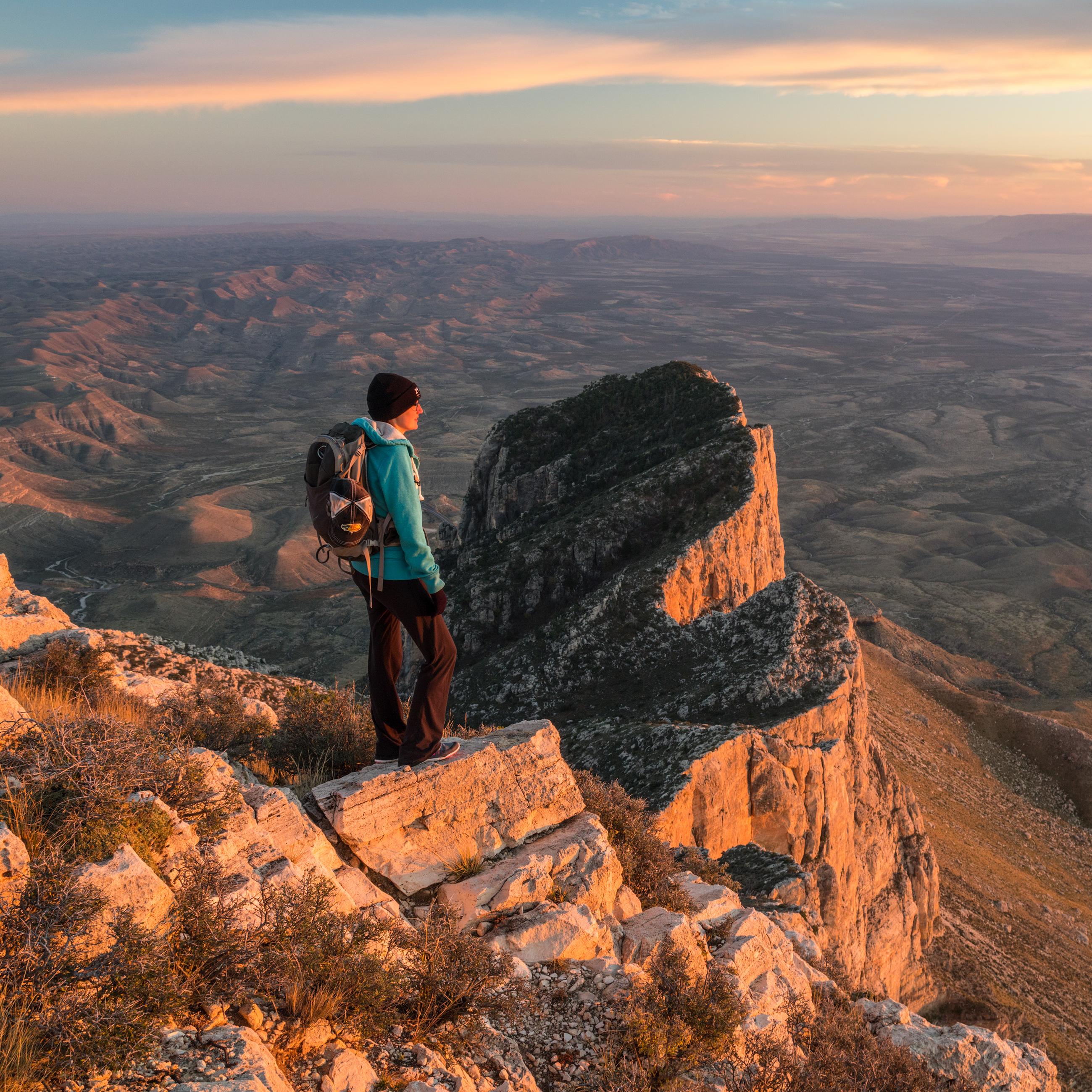 A hiker stands on a peak with lower mountains in the background at sunset