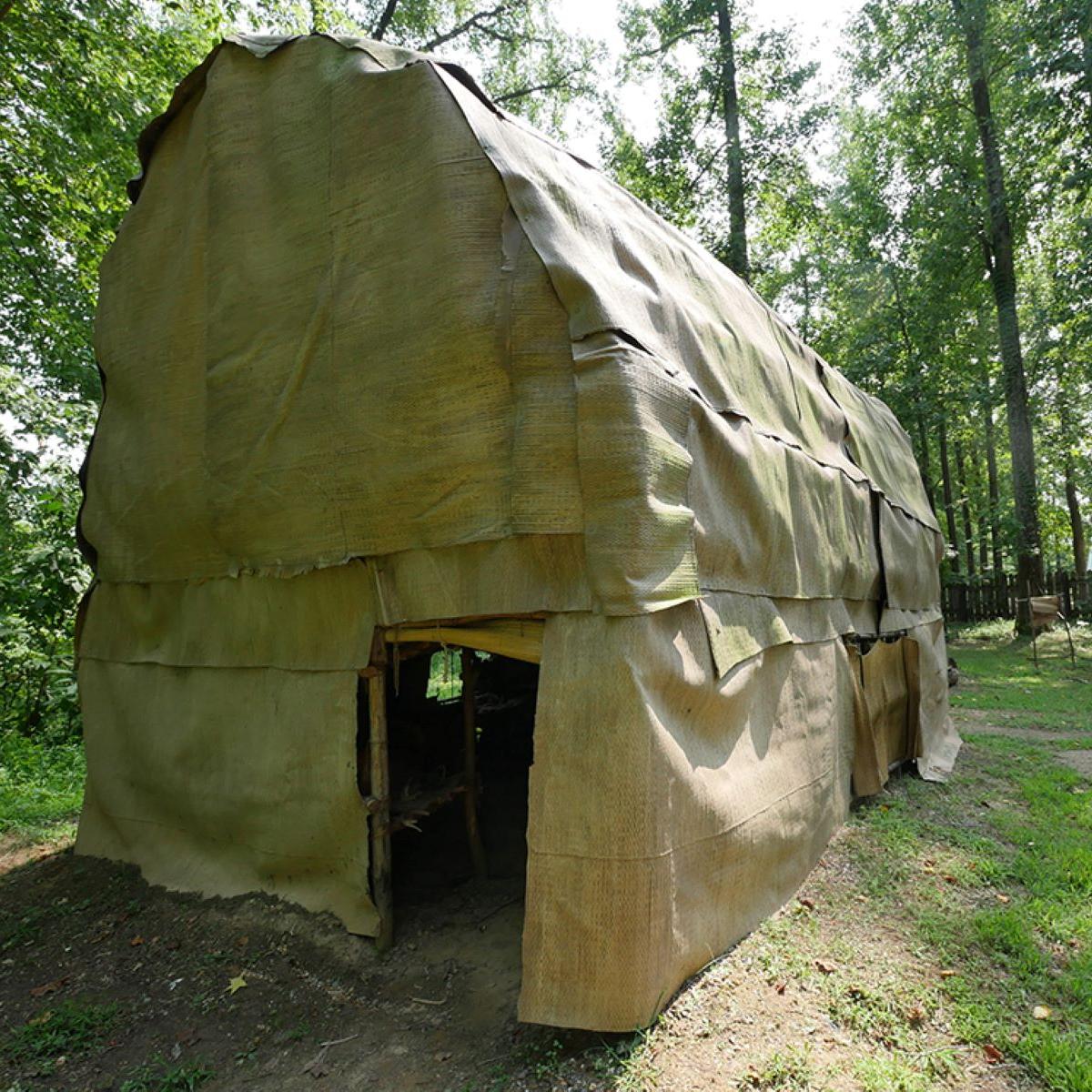 A Native American longhouse sitting in a grassy area under trees.
