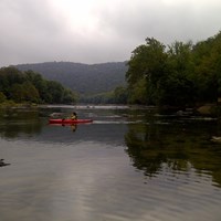 kayaker on a calm, wide river with forested hillsides on both shorelines