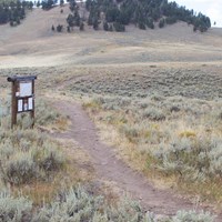 Trail heads past a wooden trailhead sign/bulletin board and out across a hilly sagebrush landscape.