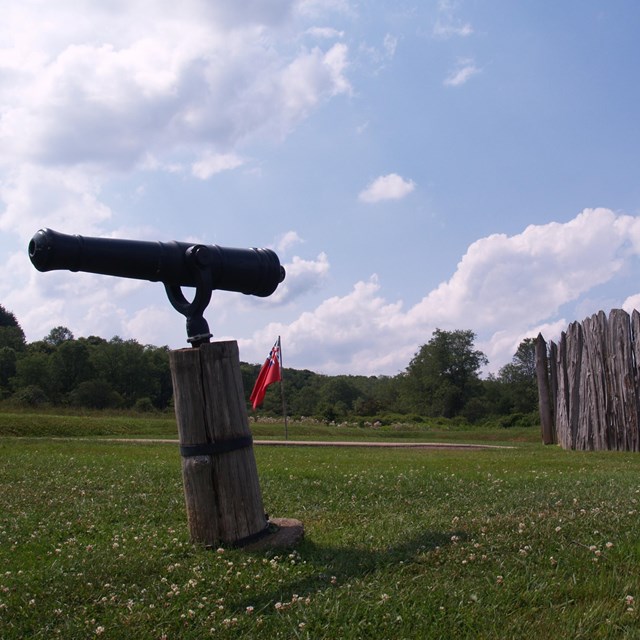 A wooden stockade in a green field with blue sky above. A red flag flutters by the stockade. 