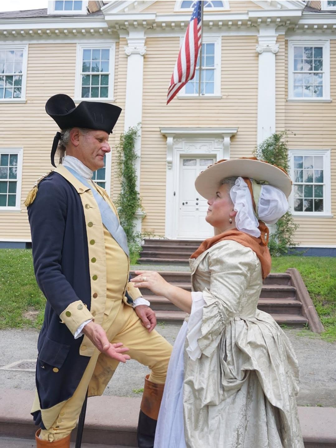 George and Martha Washington living historians holding hands facing each other in front of a house