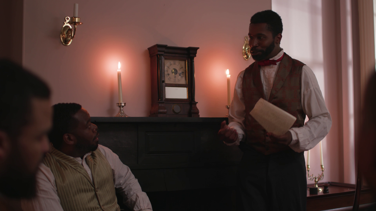 A man of African descent stands speaking to seated men in a parlor. A clock is on the mantle.