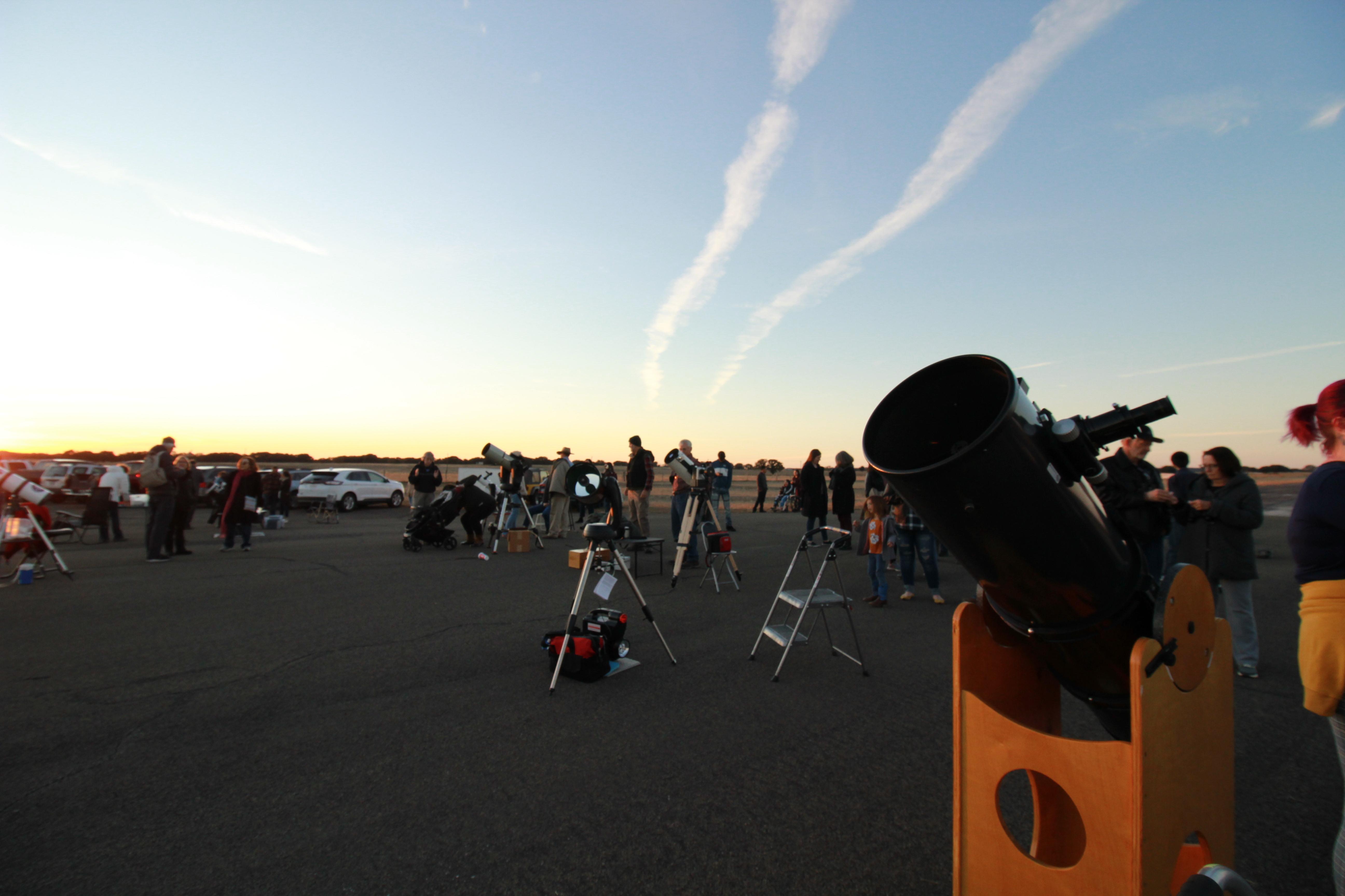 Numerous people stand next to telescopes on a wide-open asphalt area.