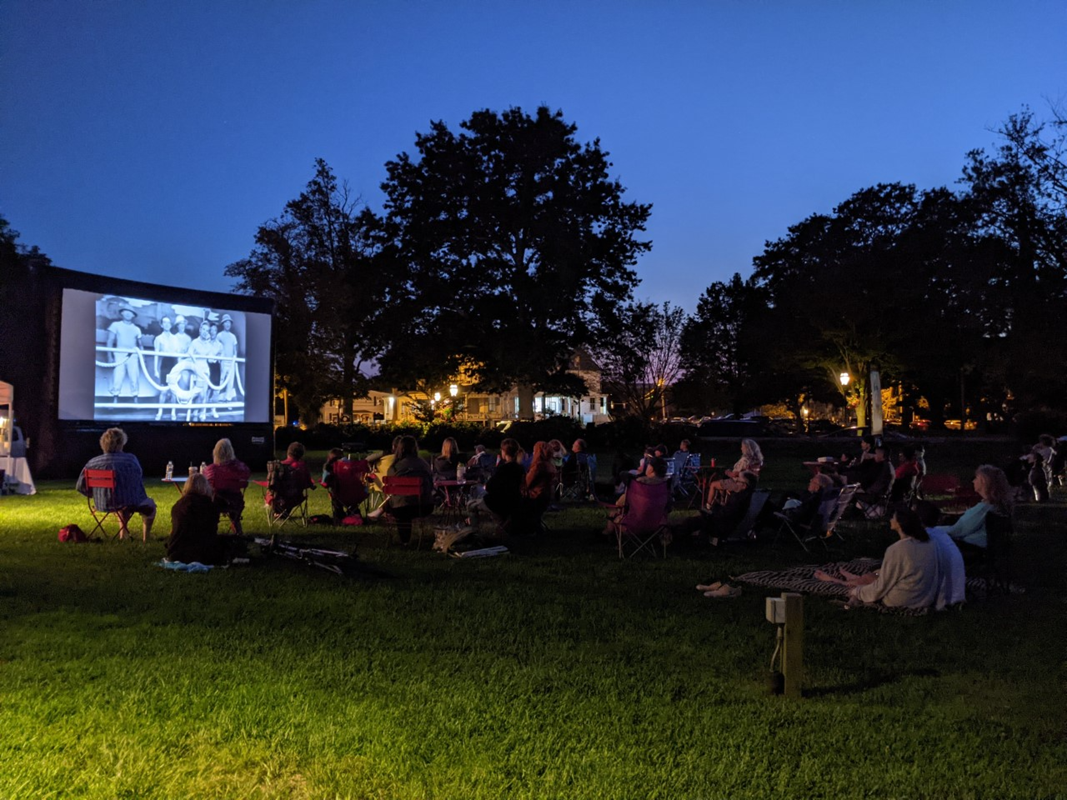 Group of people sitting on blankets and lawn chairs watch films on blow up screen