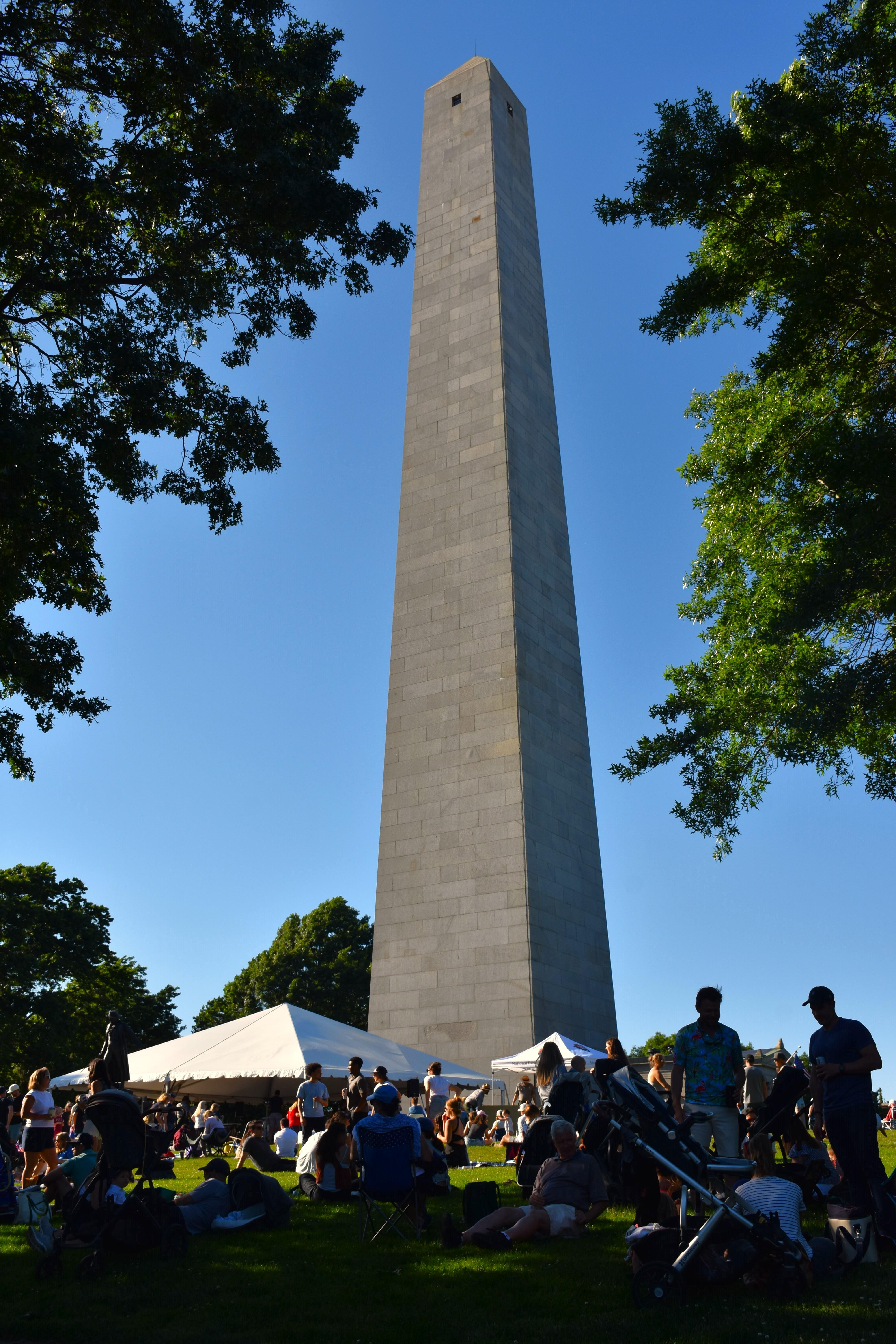 A crowd gathers on the lawn in front of the tall Bunker Hill Monument