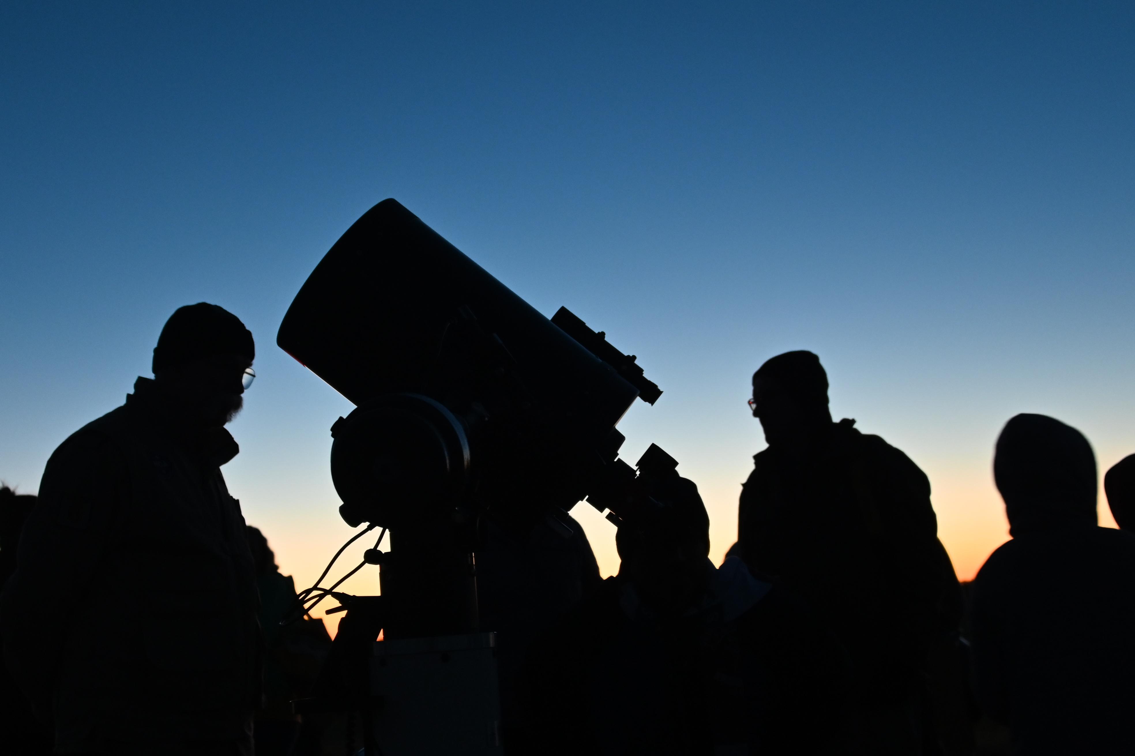 Silhouettes of people and a large telescope against the backdrop of a twilight sky.