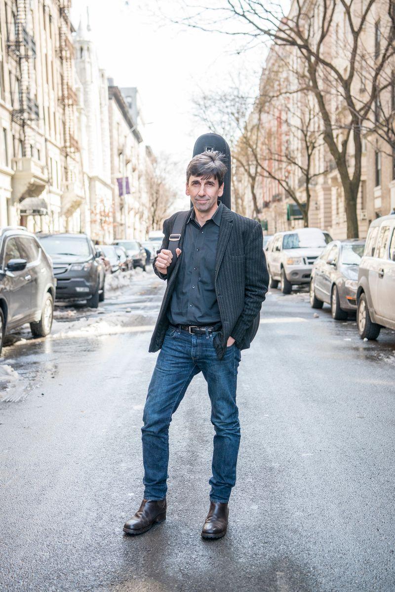 A man standing in the middle of a city street with a guitar case on his back.